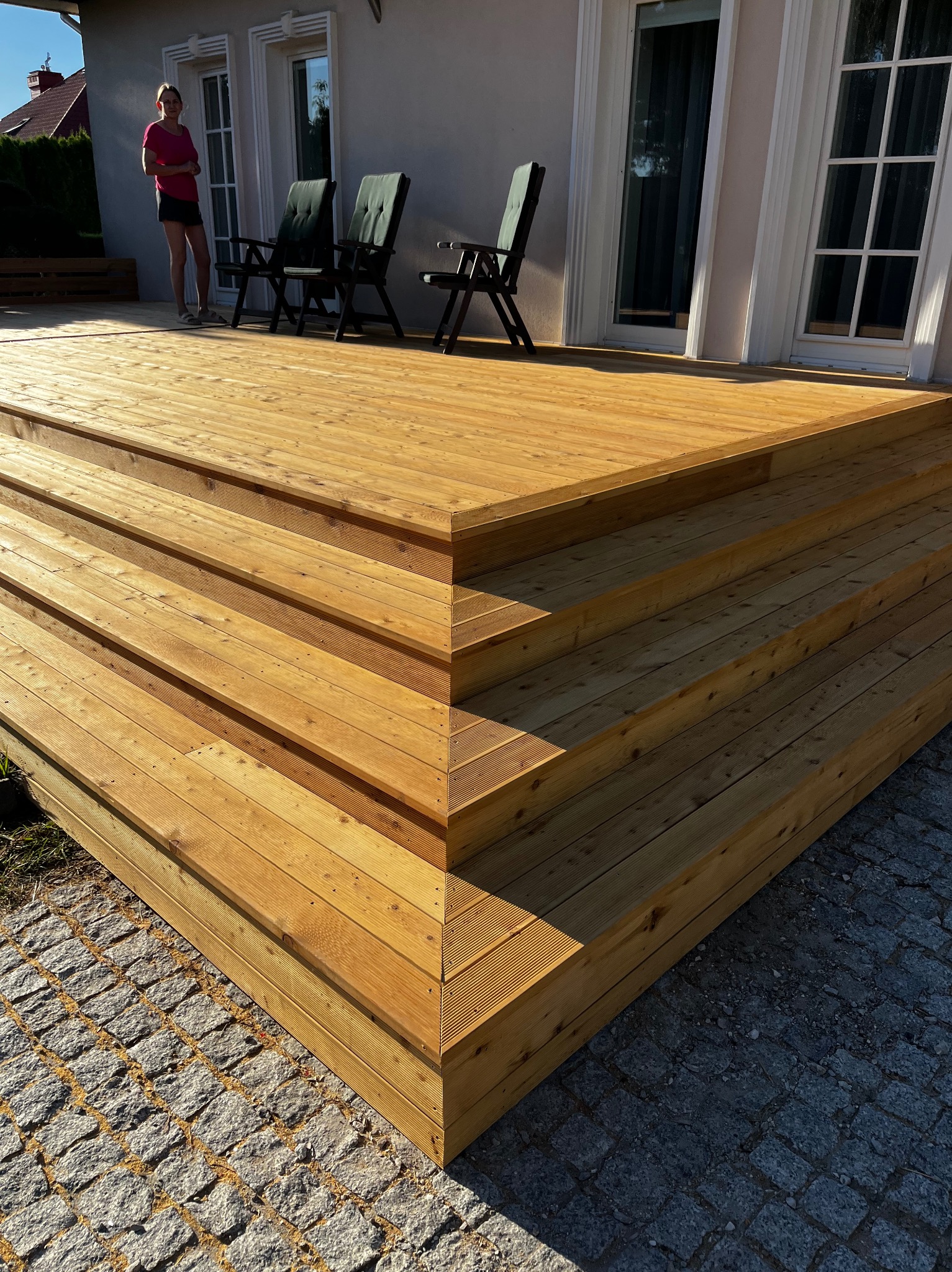 Nowy, drewniany taras schodkowy z jasnego drewna, prowadzący do drzwi balkonowych. W tle dwie osoby i krzesła ogrodowe. Kostka brukowa na pierwszym planie.