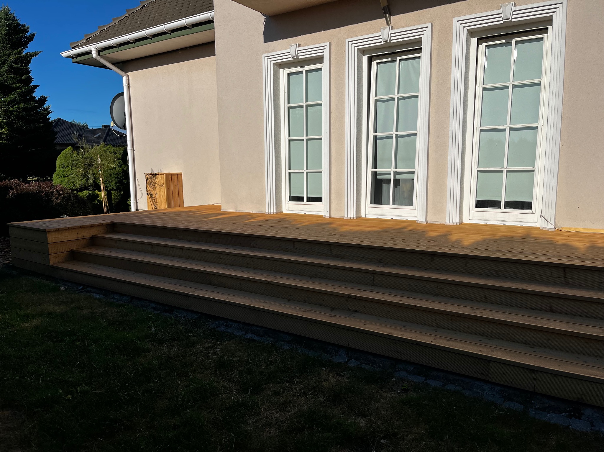 Nowy taras drewniany z czterema schodkami prowadzący do domu z trzema oknami w białych, ozdobnych ramach. Widok na fragment ogrodu.