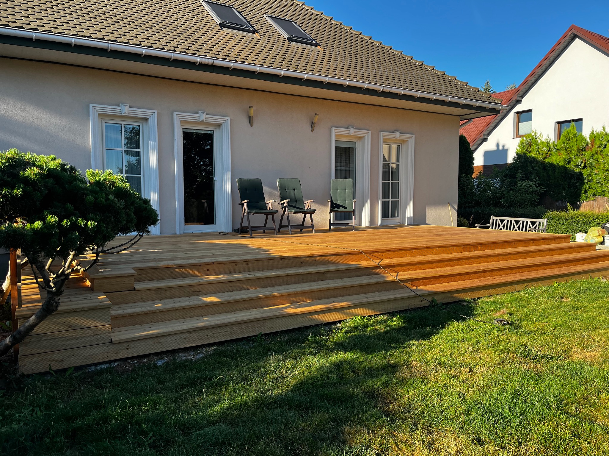 Nowoczesny taras drewniany z trzema schodkami, z widokiem na dom z oknami i krzesłami. Ujęcie w słoneczny dzień, trawnik w pierwszym planie.