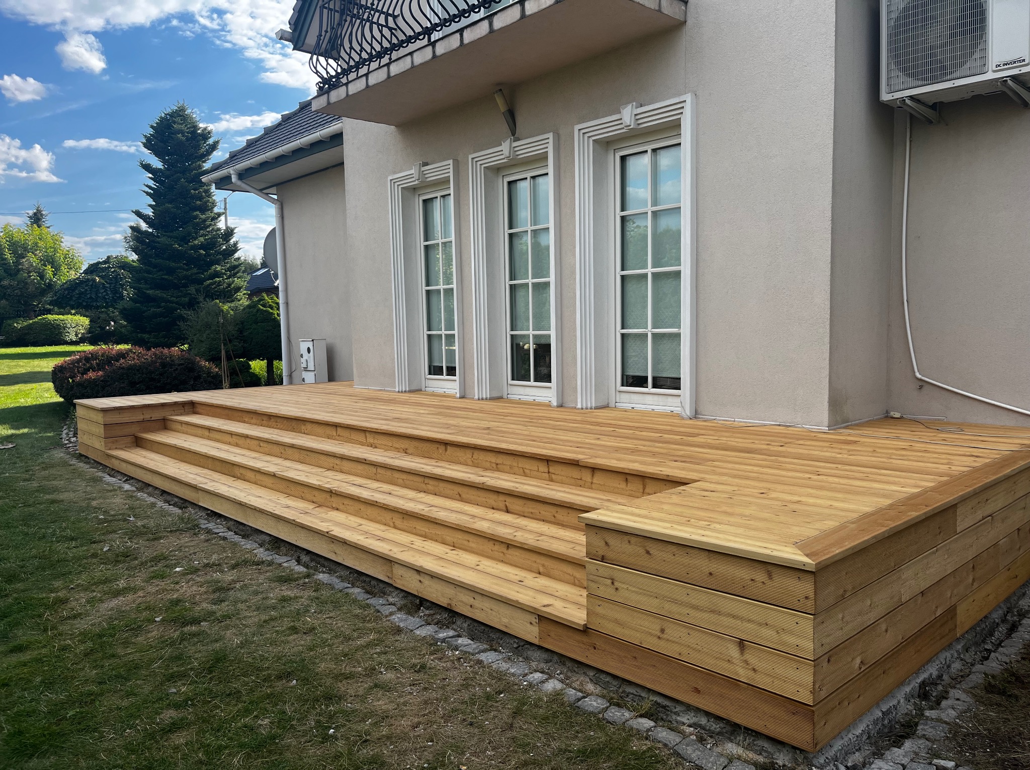 Jasny taras drewniany z niskimi schodami przylegający do budynku z trzema oknami. Widoczne elementy ogrodu i błękitne niebo.