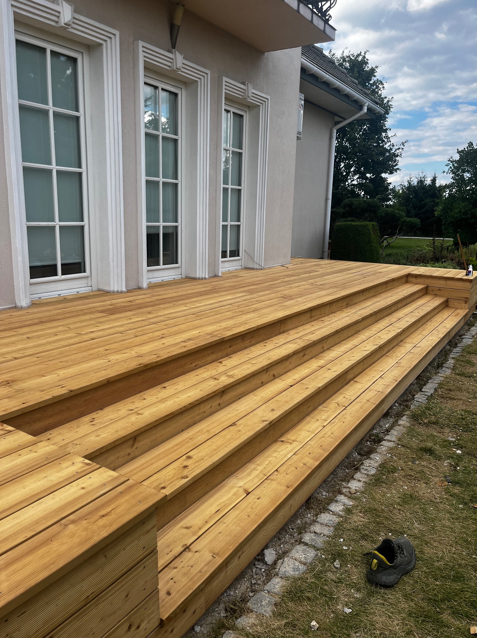 Nowy, drewniany taras z kilkoma stopniami, przylegający do budynku z białymi oknami. Na trawie leży but. Widok na ogród w tle.