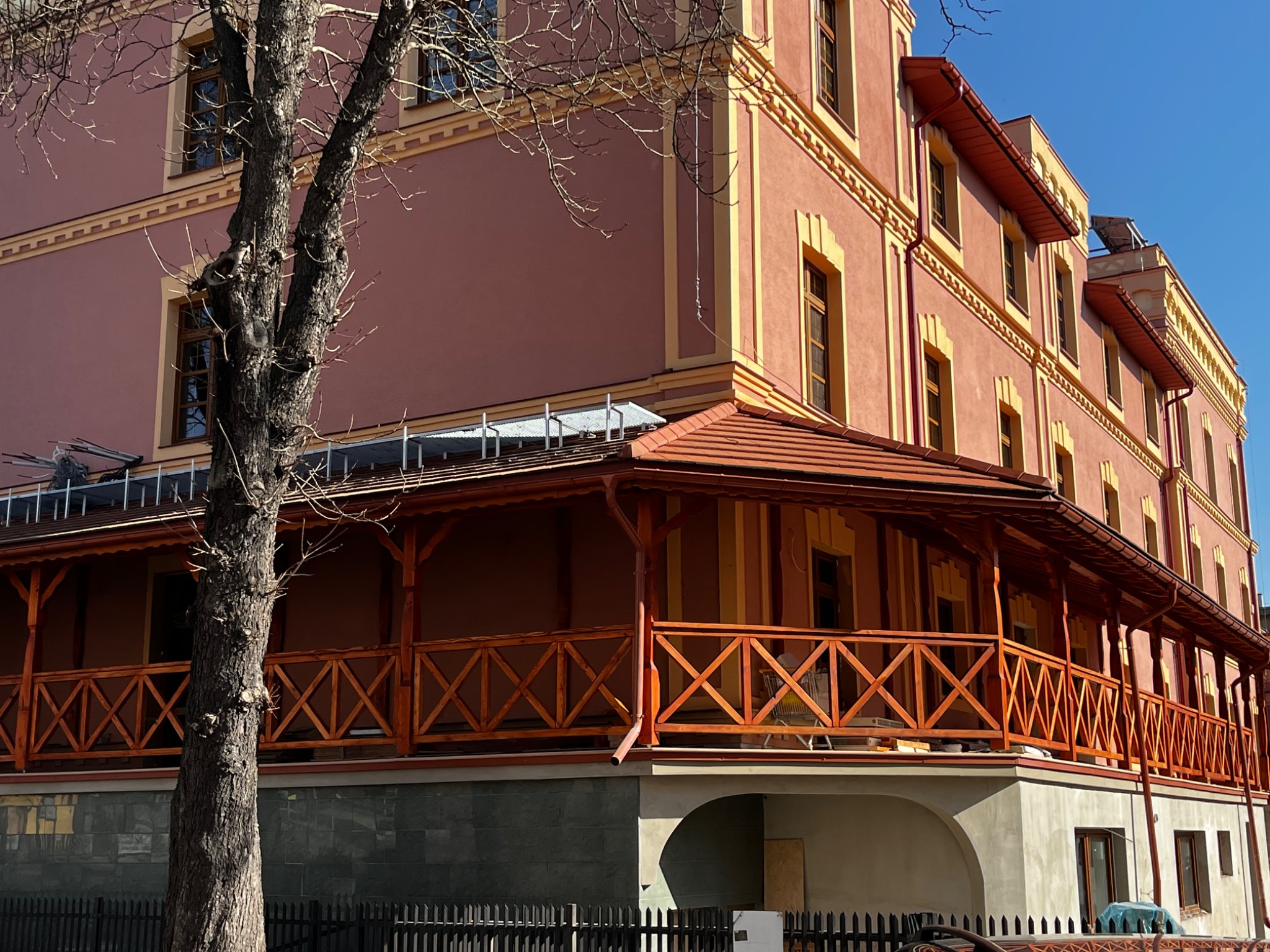 Budynek z balkonem z drewnianą balustradą w kolorze brązowym, widok z dołu, na pierwszym planie drzewo bez liści, błękitne niebo w tle.