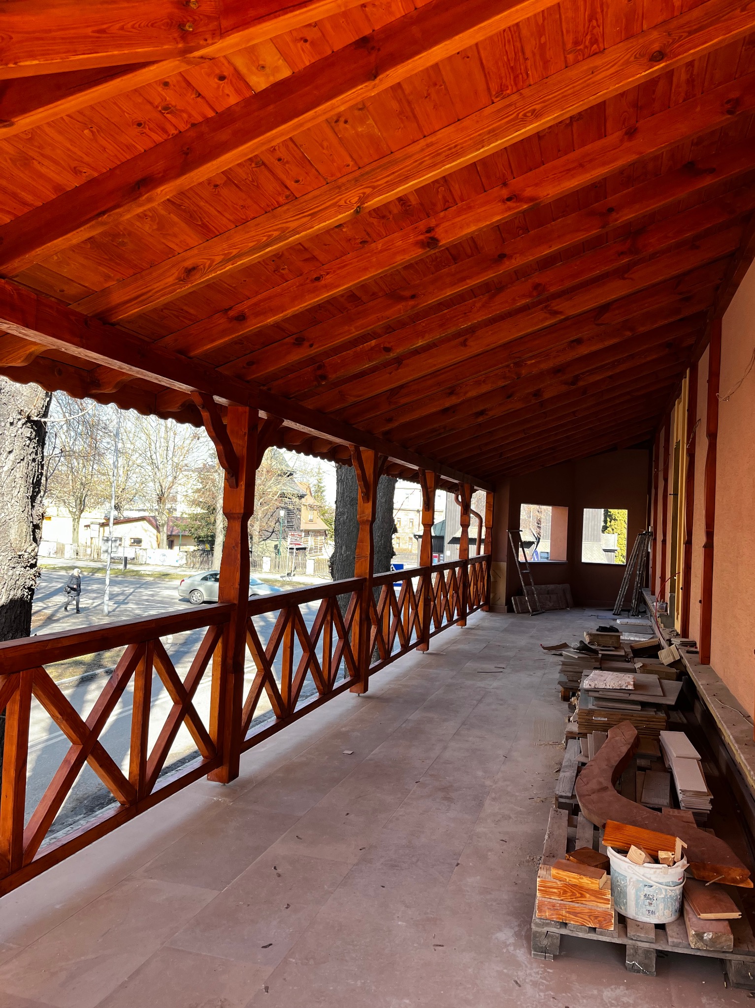 Zadaszony taras z drewnianą konstrukcją i balustradą w kolorze ciepłego brązu. Widoczne materiały budowlane i narzędzia, sugerujące etap wykończeniowy.