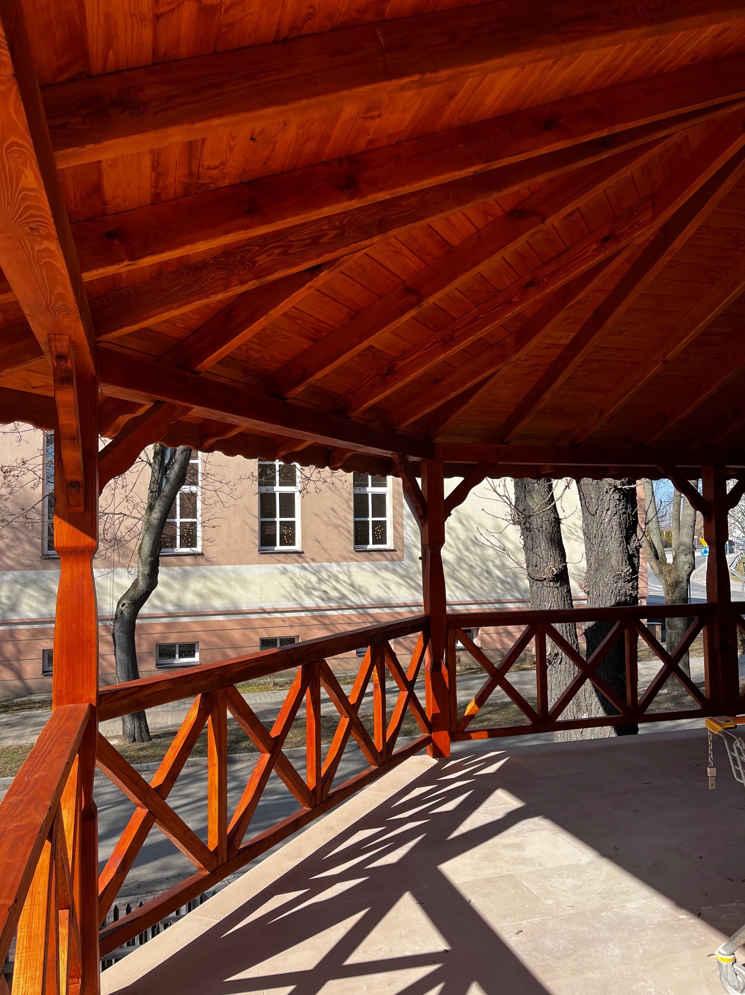 Drewniana altana z beżową podłogą w słoneczny dzień. Widoczne elementy konstrukcyjne i balustrady. W tle budynek i drzewa.