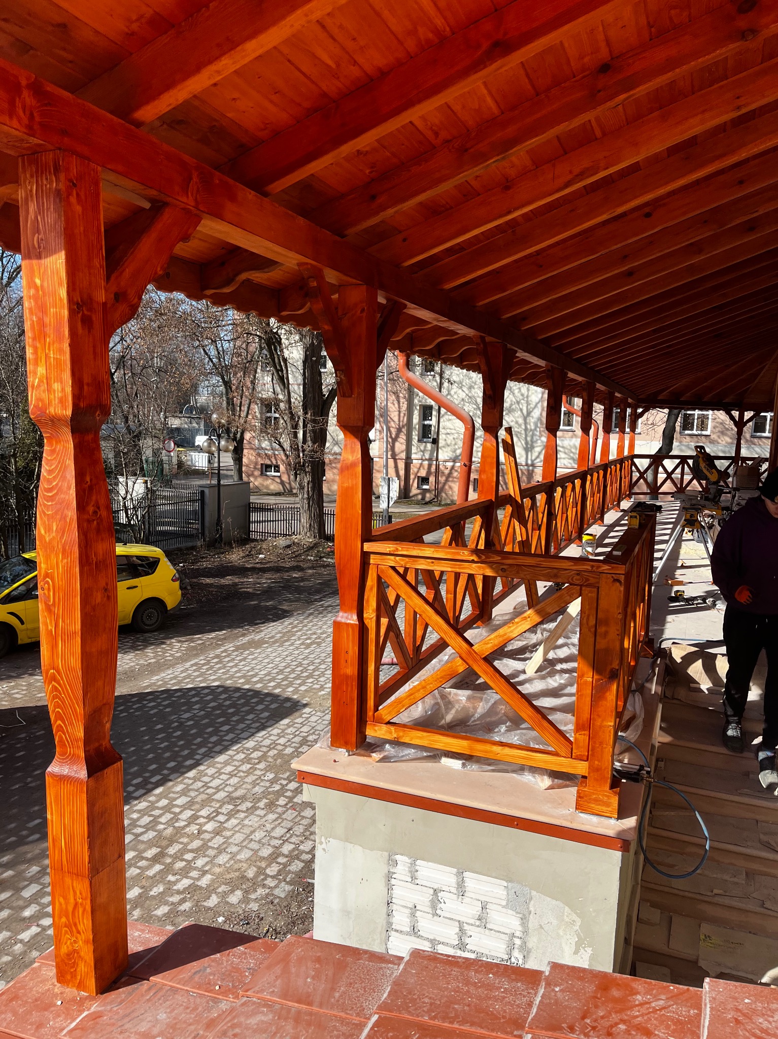 Zadaszony taras z drewnianą balustradą w kolorze miodowym, widok z zewnątrz. Częściowo wykończony, z widocznymi narzędziami i elementami konstrukcyjnymi. Żółty samochód w tle.