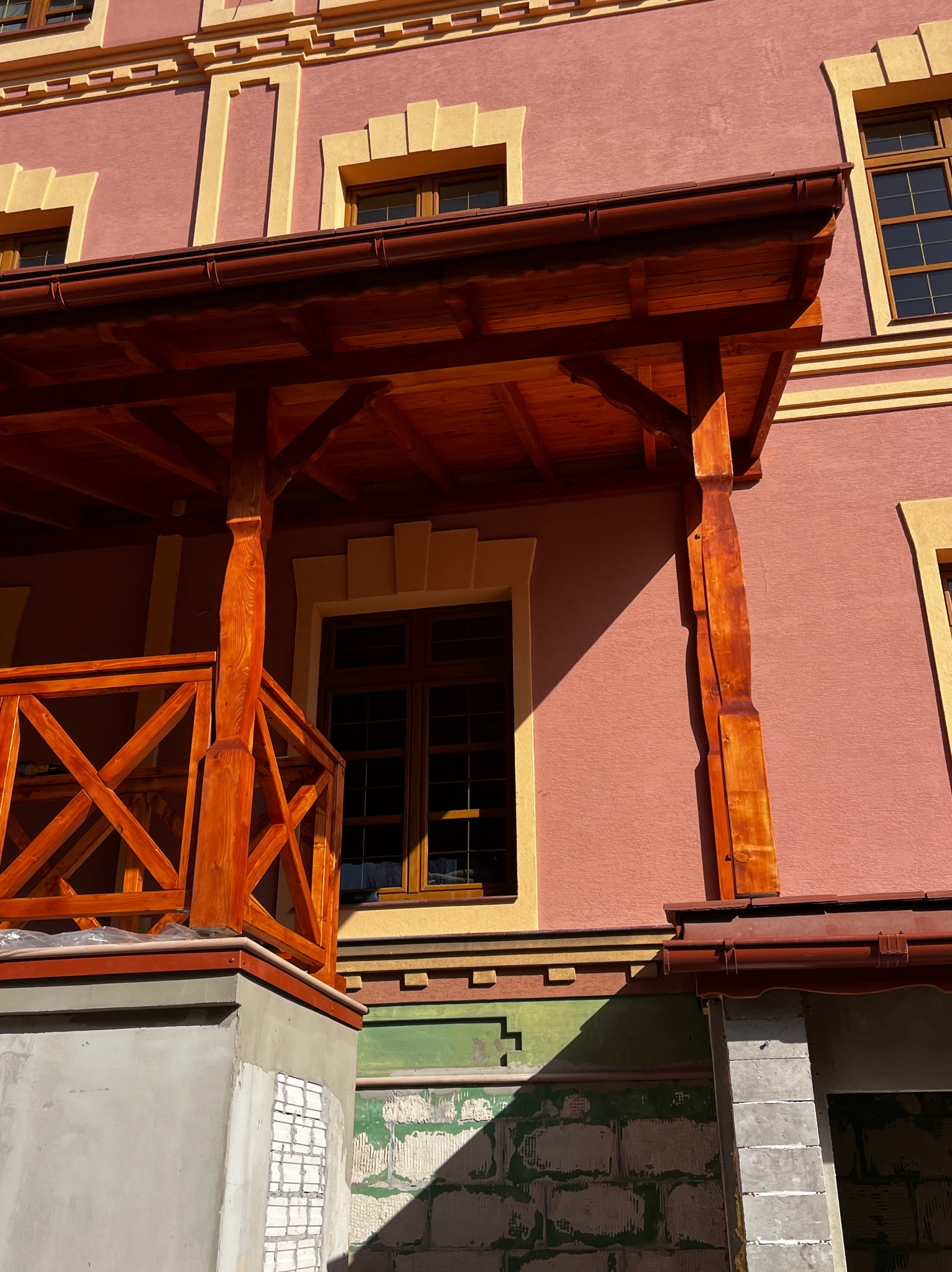 Drewniana konstrukcja z balustradą przy budynku z różową elewacją i żółtymi zdobieniami. Widoczne rynny i fragmenty niezakończonych prac murarskich.