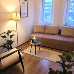 Jasny gabinet psychologiczny z beżową sofą, stolikiem z zegarem i chusteczkami, lampą podłogową, plakatem z kwiatami i roślinami doniczkowymi.