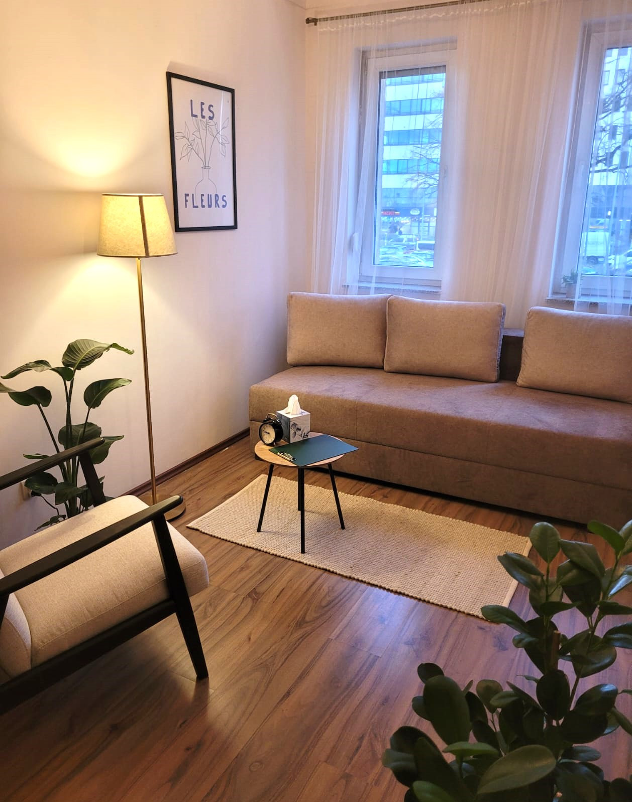 Jasny gabinet psychologiczny z beżową sofą, stolikiem z zegarem i chusteczkami, lampą podłogową, plakatem z kwiatami i roślinami doniczkowymi.