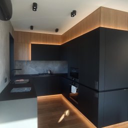 Concord Dmytro Hmyria - Nowoczesna kuchnia w czerni i drewnie z subtelnym oświetleniem LED pod szafkami, drewniana podłoga, minimalistyczne lampy sufitowe, widoczne miski dla zwierząt w rogu.