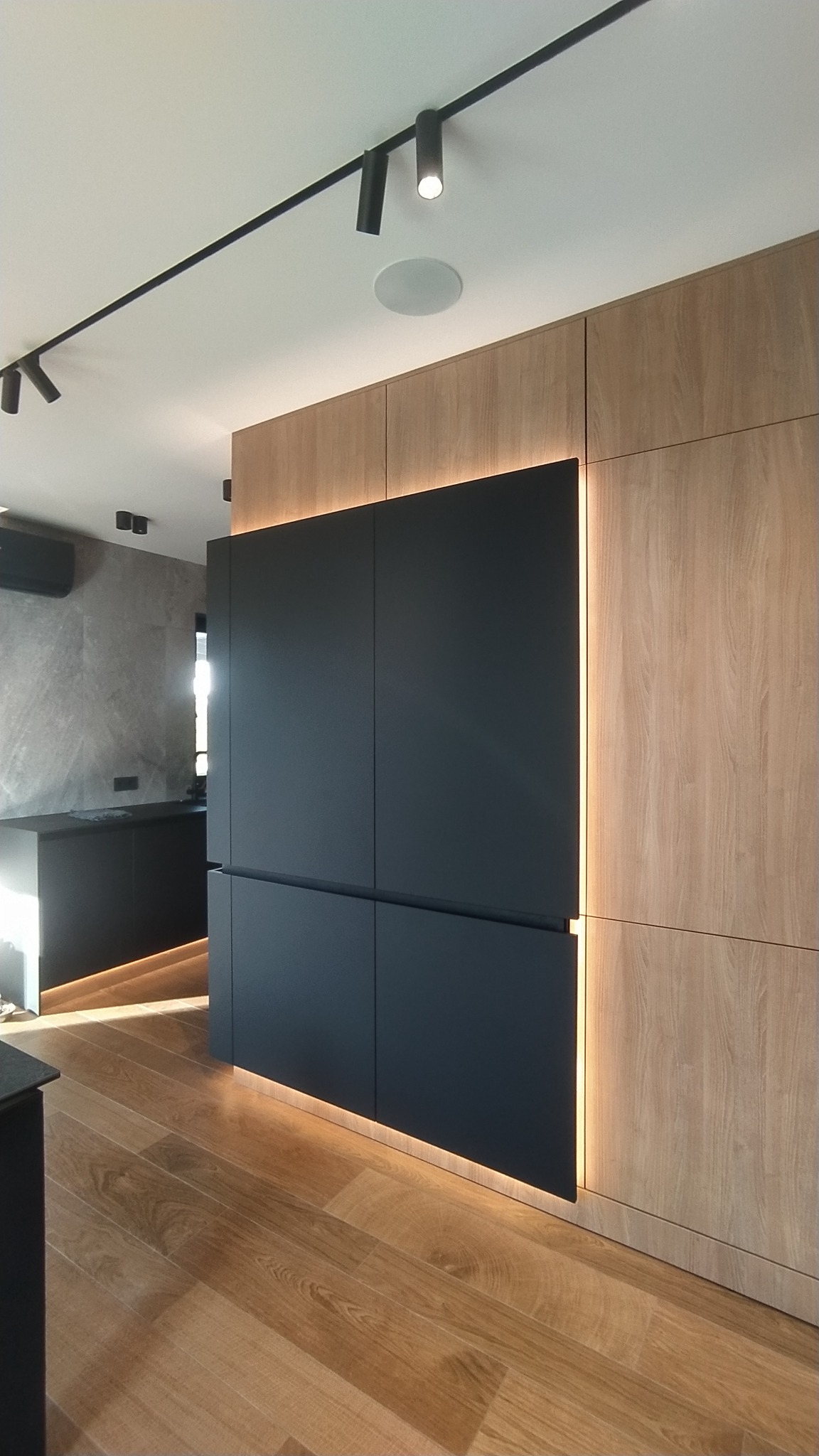 Nowoczesna zabudowa kuchenna z czarnymi frontami i drewnianymi panelami, oświetlona listwami LED, uzupełniona parkietem w jodełkę.