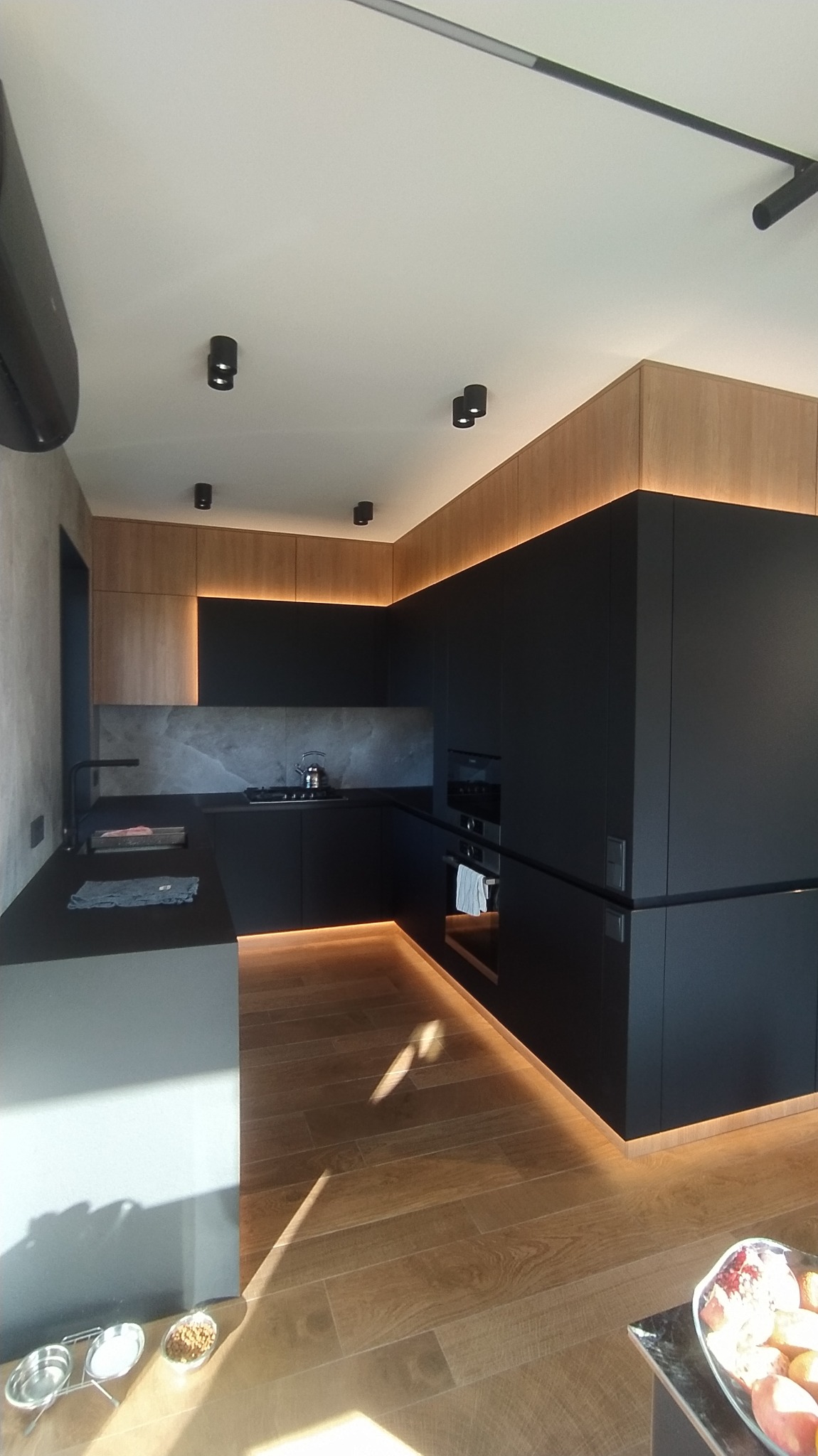 Nowoczesna kuchnia w czerni i drewnie z subtelnym oświetleniem LED pod szafkami, drewniana podłoga, minimalistyczne lampy sufitowe, widoczne miski dla zwierząt w rogu.