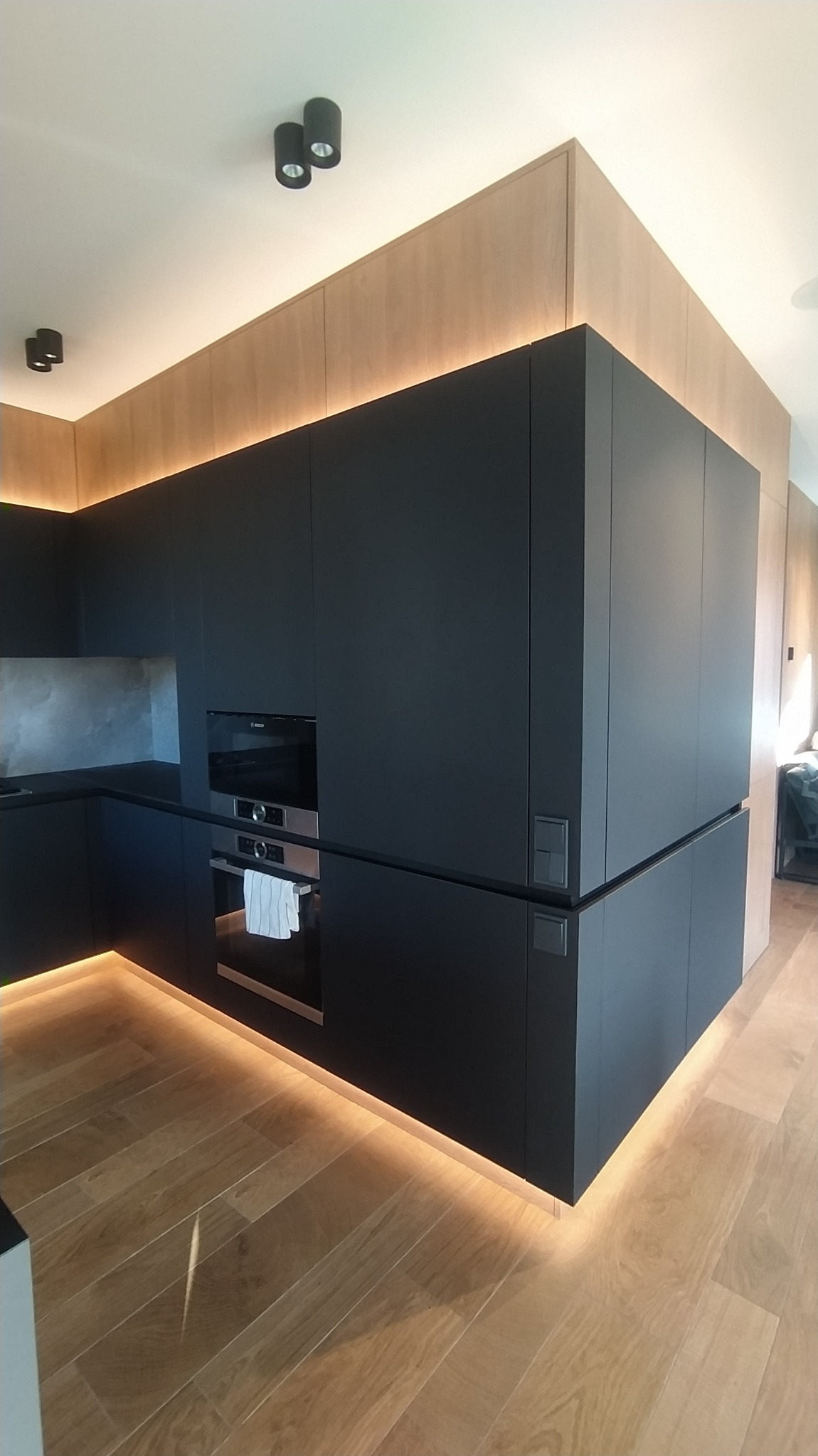 Nowoczesna kuchnia z czarnymi matowymi frontami, podświetleniem LED pod szafkami i na górze, drewniana podłoga i okładzina ścienna, minimalistyczne oświetlenie punktowe.