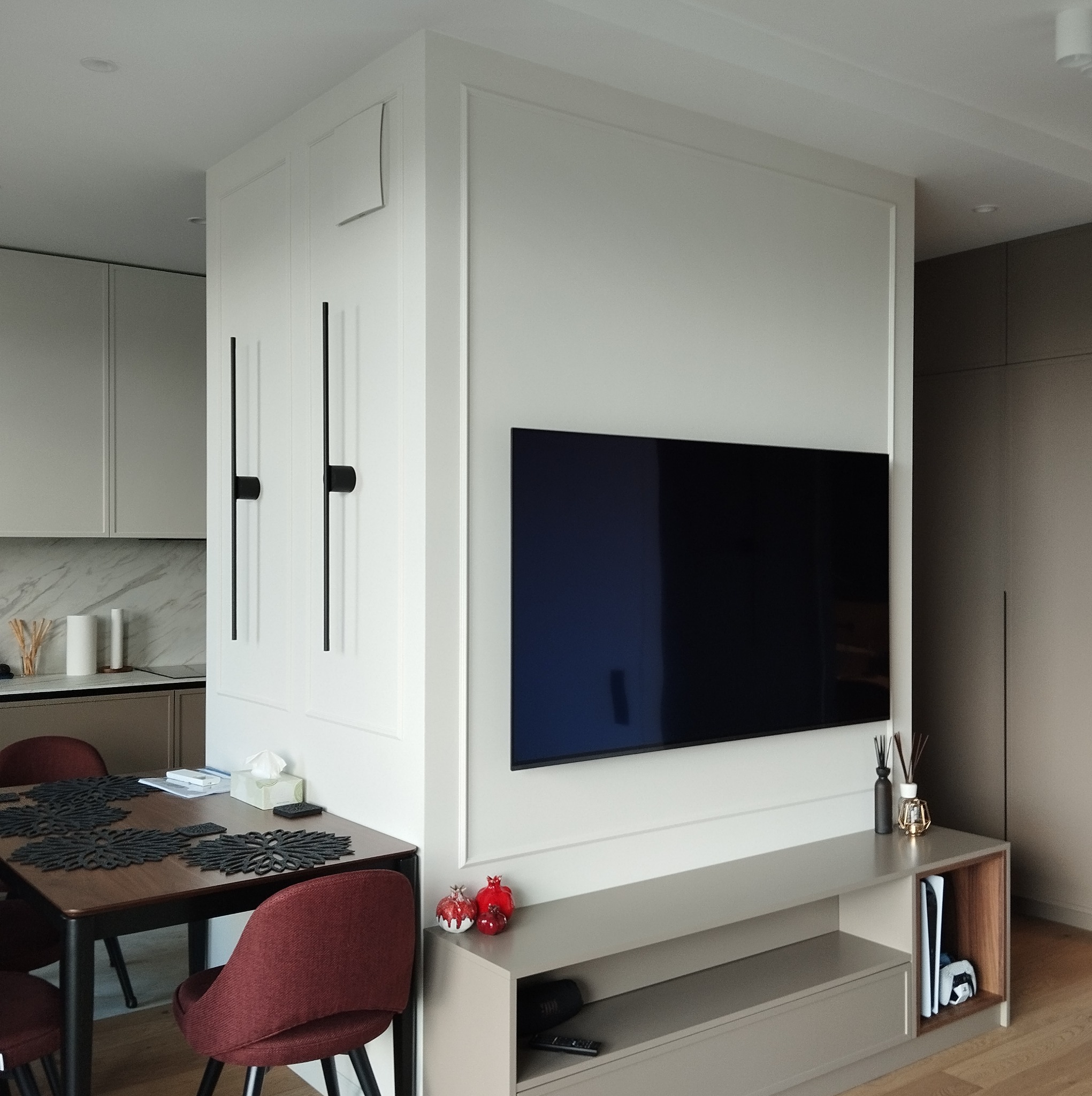 Salon z telewizorem na ścianie, minimalistyczna szafka RTV, stół z bordowymi krzesłami, nowoczesne oświetlenie ścienne i dekoracje w postaci ceramicznych granatów.