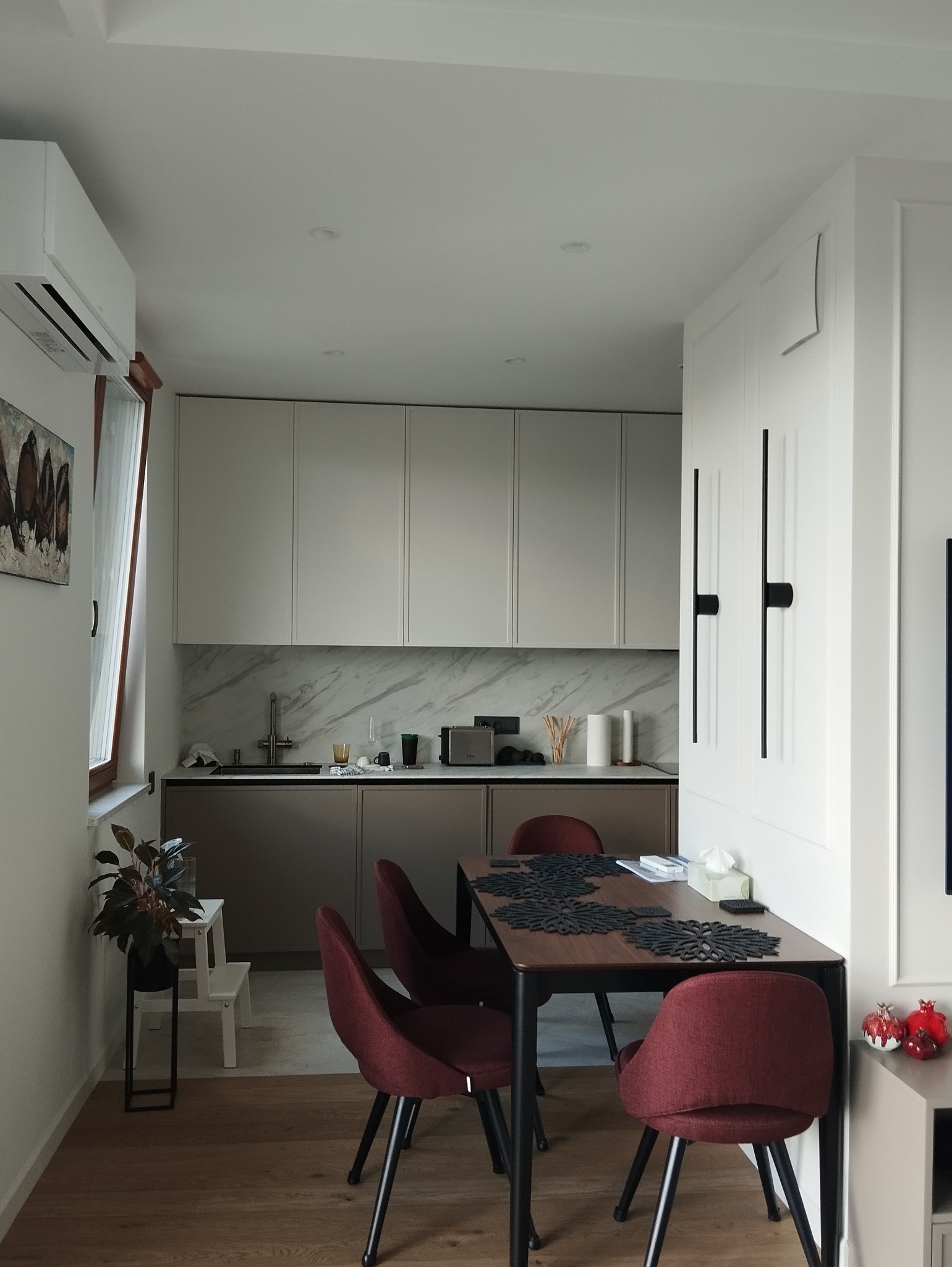 Nowoczesna kuchnia z jadalnią: beżowe szafki górne, brązowe dolne, marmurowy backsplash, stół z czterema bordowymi krzesłami i dekoracyjnymi podkładkami.