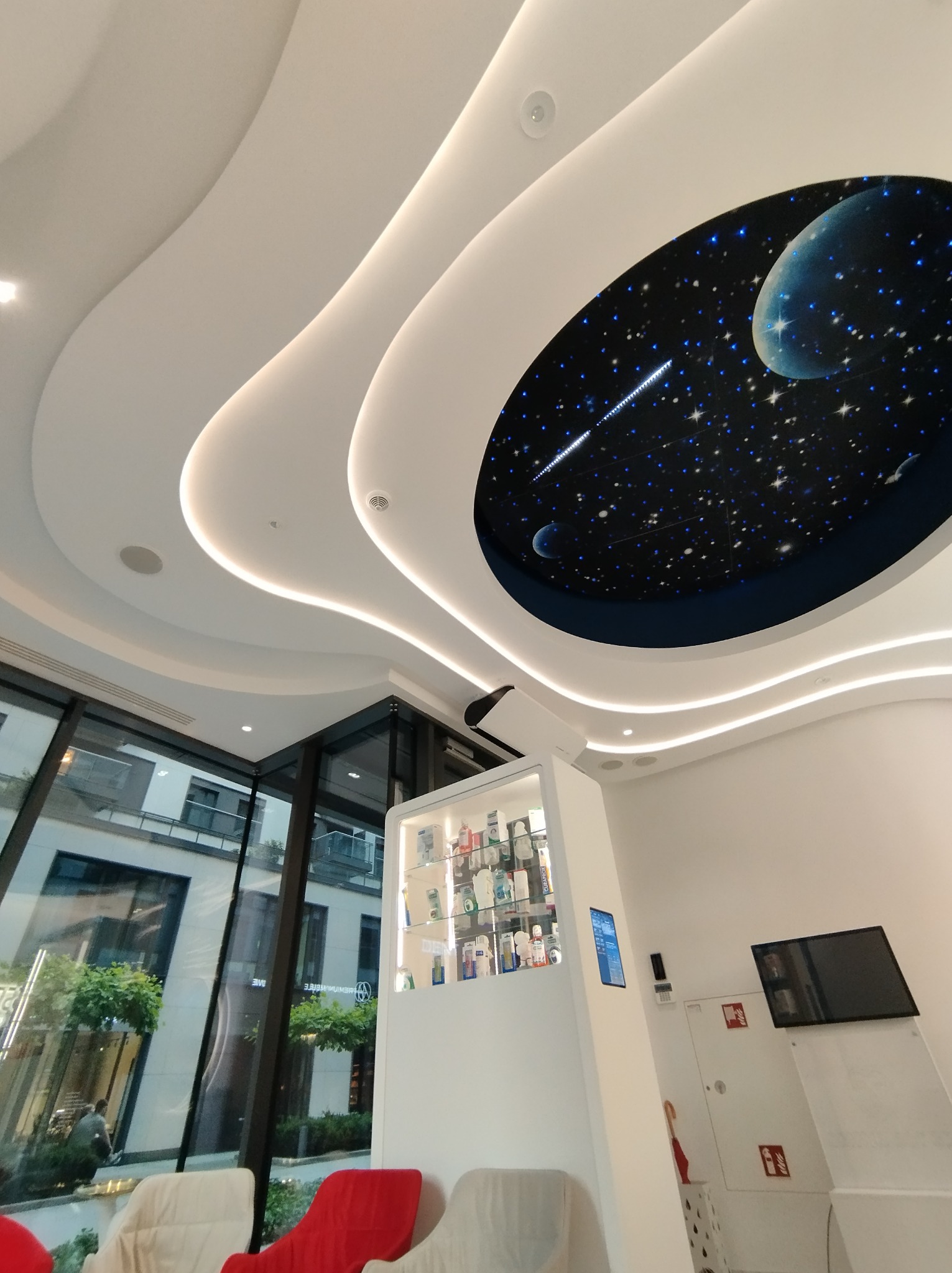 Nowoczesne wnętrze biura z falistym sufitem podwieszanym i oświetleniem LED, w centralnej części sufitu dekoracja imitująca kosmos z planetami i gwiazdami, przeszklona ściana z widokiem na zieleń.