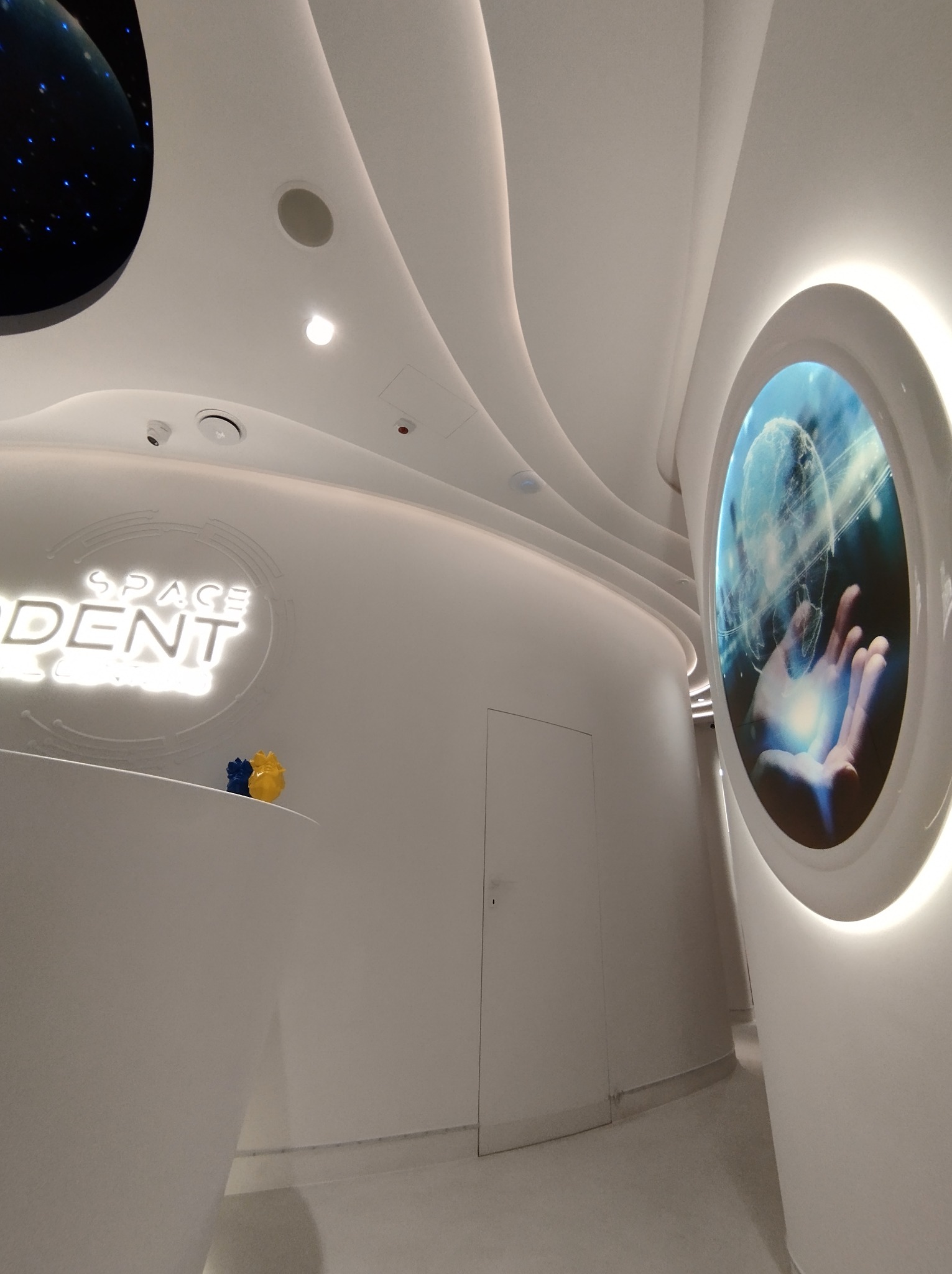 Nowoczesne białe wnętrze biura z futurystycznym designem, zaokrąglone ściany i sufit, logo firmy 'SPACE DENT' podświetlone, dekoracyjny okrągły ekran z grafiką dłoni trzymającej globus...