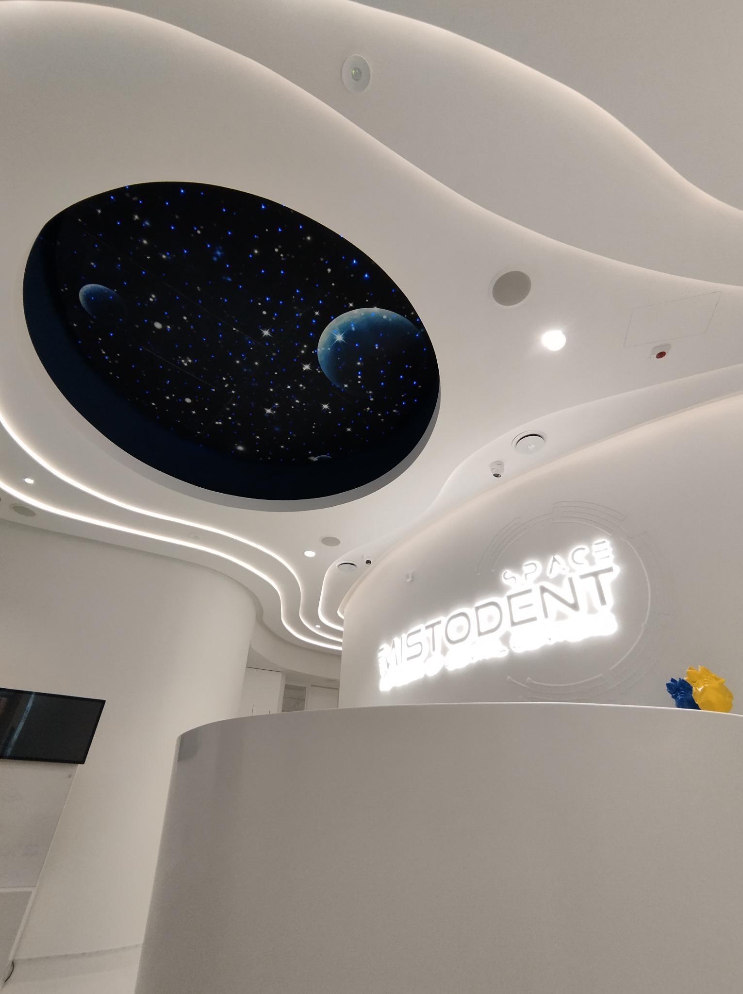 Recepcja w nowoczesnym biurze z podświetlanym logo 'MISTODENT SPACE' i sufitem z motywem kosmosu, oświetlenie LED wzdłuż zakrzywionych ścian.