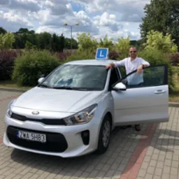 Instruktor opiera się o srebrny samochód z otwartymi drzwiami, na dachu znak 'L', w tle zieleń i błękitne niebo.