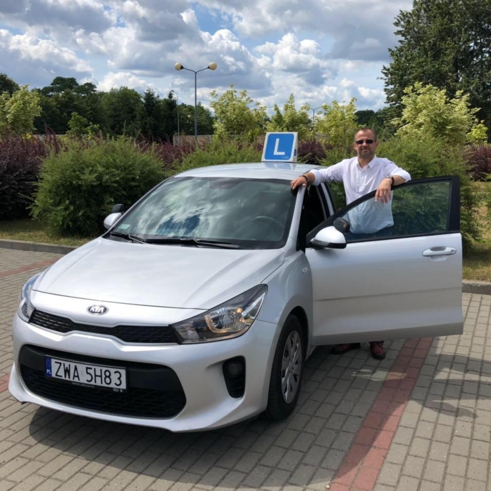 Instruktor opiera się o srebrny samochód z otwartymi drzwiami, na dachu znak 'L', w tle zieleń i błękitne niebo.