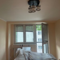 Mateusz Rybarski - Pokój w trakcie remontu: ściany pomalowane na beżowo, okno z żaluzjami, grzejnik i podłoga przykryte folią ochronną, sufit z nowoczesną lampą.