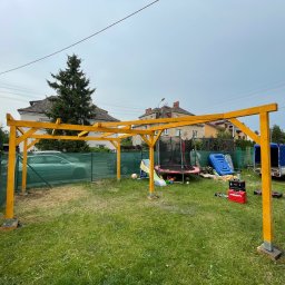 Mateusz Rybarski - Konstrukcja drewnianej altany w trakcie budowy, żółte belki na metalowych podstawach, trawnik z zabawkami w tle, widoczny samochód i przyczepa.