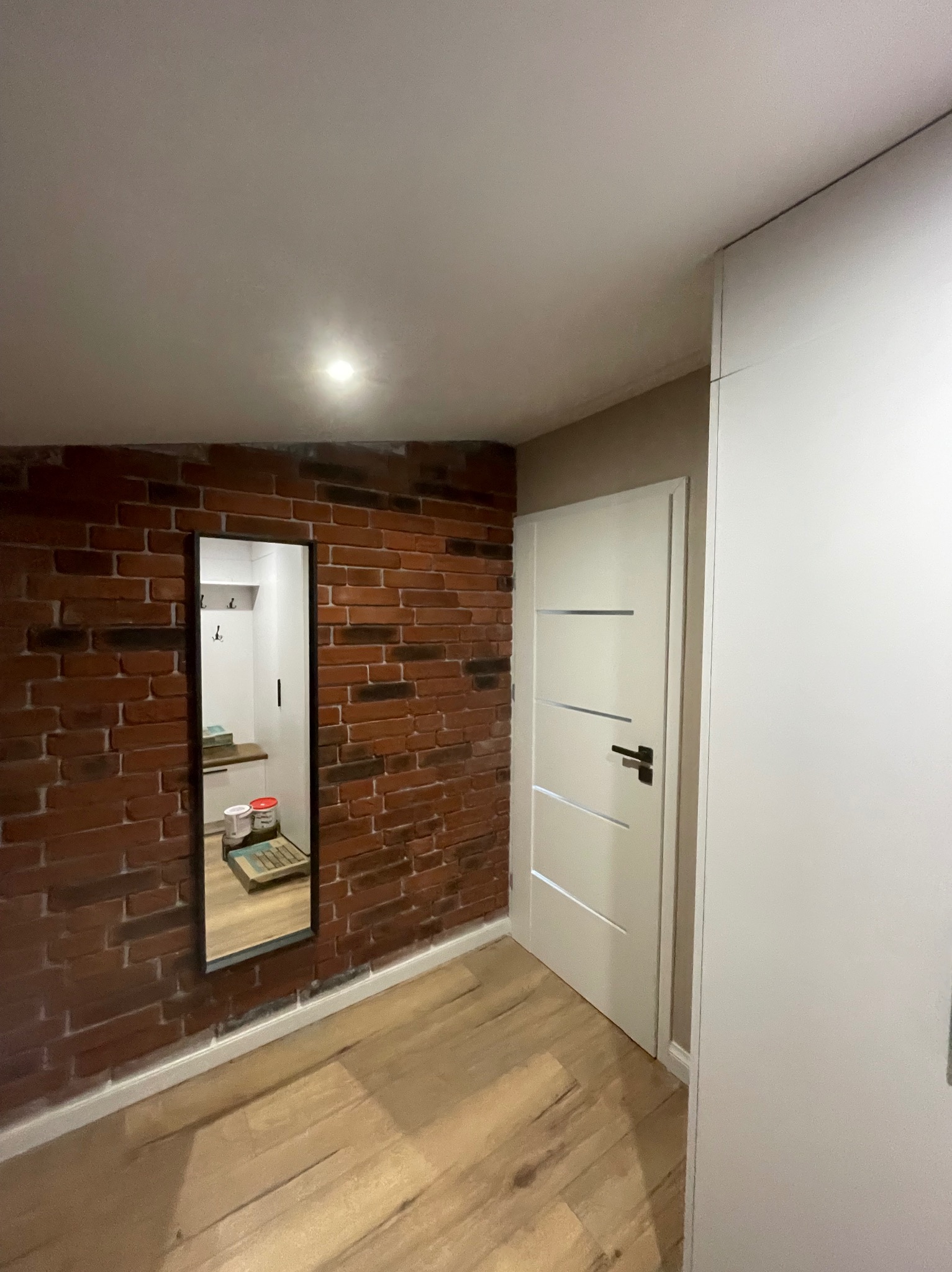 Wnętrze po remoncie z ceglaną ścianą, białymi drzwiami i szafą wnękową; w lustrze odbijają się puszki z farbą i akcesoria malarskie.