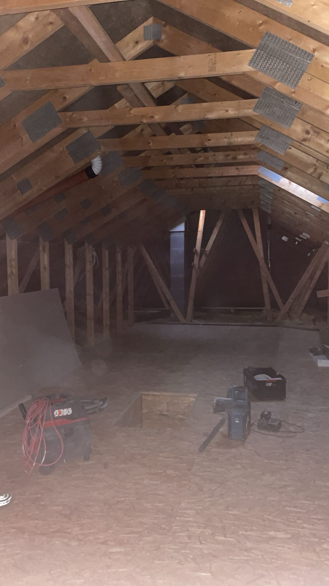 Surowe poddasze z drewnianą konstrukcją dachu, widoczne narzędzia i materiały budowlane, w tym odkurzacz przemysłowy i poziomica laserowa na trójnogu, przygotowane do dalszych prac wykończeniowych.