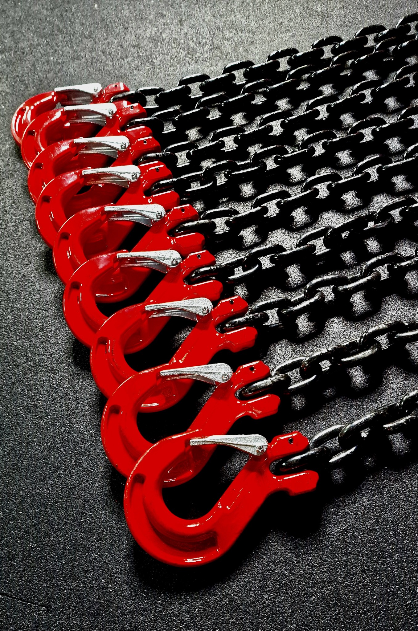 Rząd czerwonych haków z zabezpieczeniami połączonych z czarnym łańcuchem na ciemnym, teksturowanym tle.