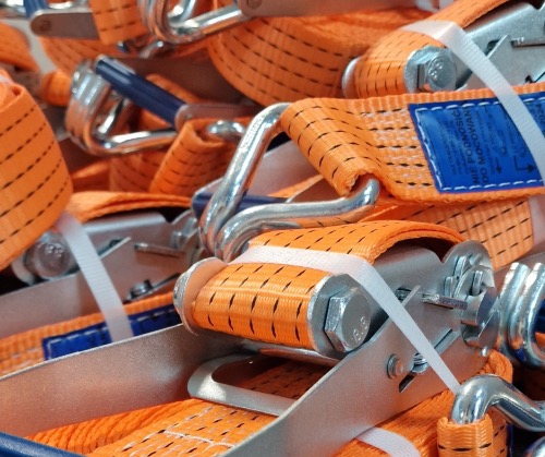 Pomarańczowe pasy transportowe z metalowymi elementami mocującymi, ułożone warstwami, widoczne białe taśmy zabezpieczające.