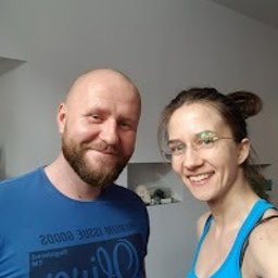 Fizjofaza Gdańsk - fizjoterapia - inhalacja wodorowa - masaż - Uśmiechnięty mężczyzna z brodą w niebieskiej koszulce stoi obok uśmiechniętej kobiety w okularach i niebieskim topie na tle jasnego wnętrza.