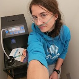 Fizjofaza Gdańsk - fizjoterapia - inhalacja wodorowa - masaż - Kobieta w okularach i bluzie z kapturem, z kaniulą nosową, robiąca selfie na tle komory hiperbarycznej z ulotką informacyjną.