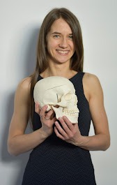 Kobieta w czarnej sukience trzyma model ludzkiej czaszki na tle białej ściany, prezentując go z uśmiechem.