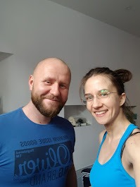 Uśmiechnięty mężczyzna z brodą w niebieskiej koszulce stoi obok uśmiechniętej kobiety w okularach i niebieskim topie na tle jasnego wnętrza.