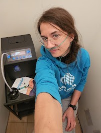 Kobieta w okularach i bluzie z kapturem, z kaniulą nosową, robiąca selfie na tle komory hiperbarycznej z ulotką informacyjną.