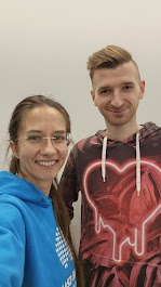 Kobieta w okularach i błękitnej bluzie oraz mężczyzna w bluzie z kapturem z motywem serca, pozujący do selfie na tle białej ściany.