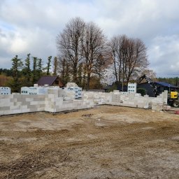 MDM COUNSTRUCTION SPÓŁKA Z OGRANICZONĄ ODPOWIEDZIALNOŚCIĄ - Wznoszenie ścian parteru domu z bloczków H+H. Na paletach składowane bloczki. W tle inne domy i drzewa. Żółta koparka stoi obok budowy. Pochmurne niebo.