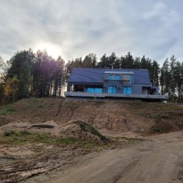 MDM COUNSTRUCTION SPÓŁKA Z OGRANICZONĄ ODPOWIEDZIALNOŚCIĄ - Surowy dom z niebieskimi oknami na wzgórzu, otoczony lasem iglastym w jesiennej scenerii. Widoczny teren budowy i droga dojazdowa.