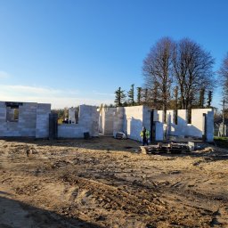 MDM COUNSTRUCTION SPÓŁKA Z OGRANICZONĄ ODPOWIEDZIALNOŚCIĄ - Wznoszenie ścian parteru z betonu komórkowego, widoczny pracownik w odblaskowej kamizelce i palety z bloczkami. Budowa w toku, słoneczny dzień.