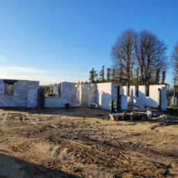 Wznoszenie ścian parteru z betonu komórkowego, widoczny pracownik w odblaskowej kamizelce i palety z bloczkami. Budowa w toku, słoneczny dzień.