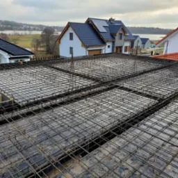 Zbrojenie stropu na budowie domu jednorodzinnego z widokiem na jezioro w tle, pochmurny dzień. Siatka z prętów stalowych ułożona na szalunku.