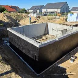 Zabezpieczony, betonowy fundament w wykopie, z wystającymi prętami zbrojeniowymi, na tle domów jednorodzinnych i pracującego robotnika. Widoczne materiały izolacyjne.