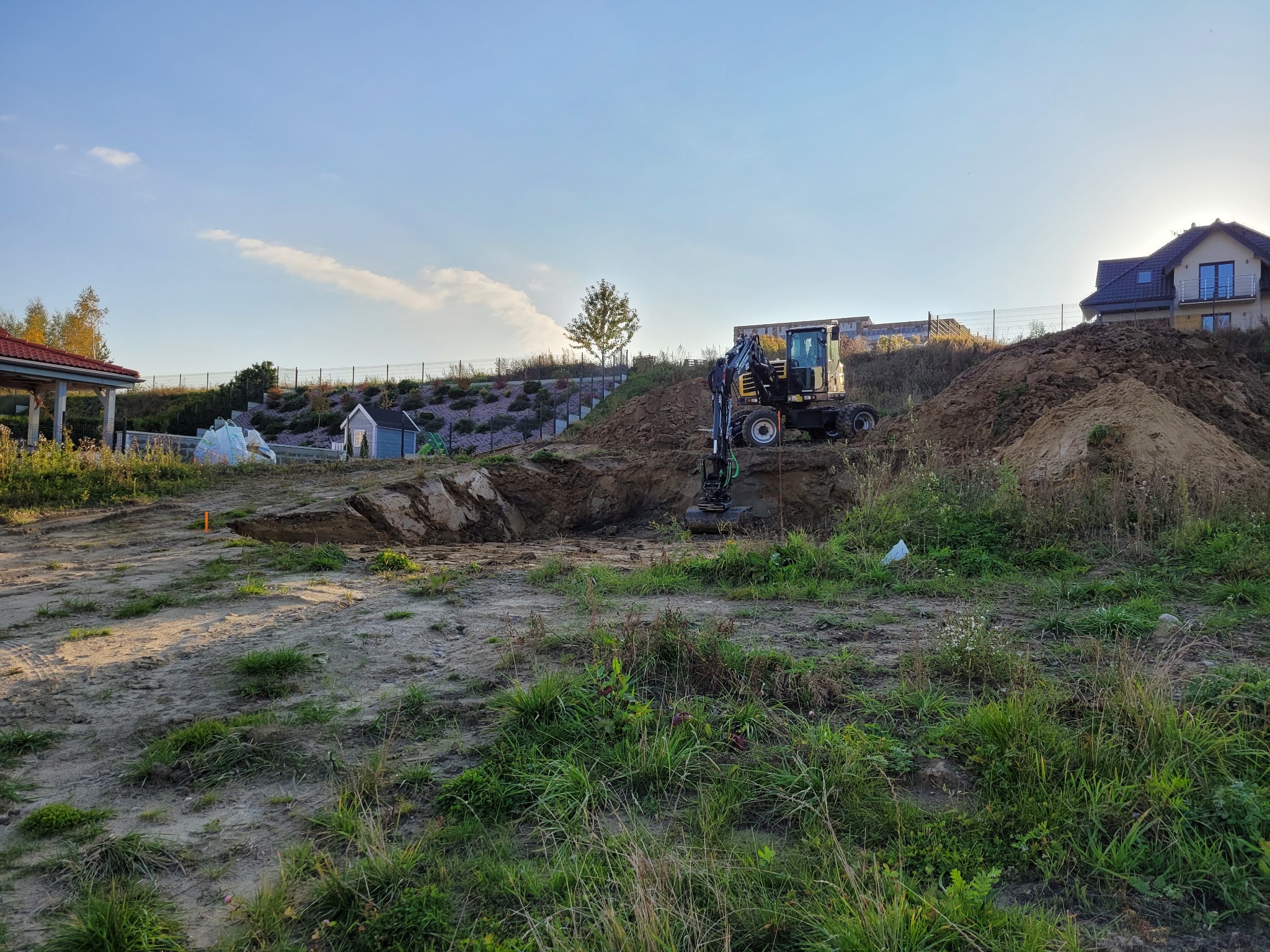 Mała koparka na placu budowy, przygotowuje teren pod fundamenty domu jednorodzinnego. W tle widoczny dom w budowie i zielone wzgórze.