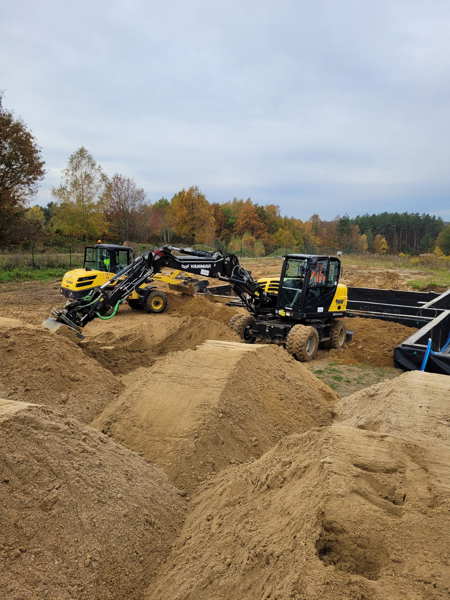 Żółte koparki Yanmar w Łężycach pracują przy wykopie pod fundamenty. W tle drzewa z jesiennymi liśćmi i szare niebo. Widoczne czarne elementy szalunków.