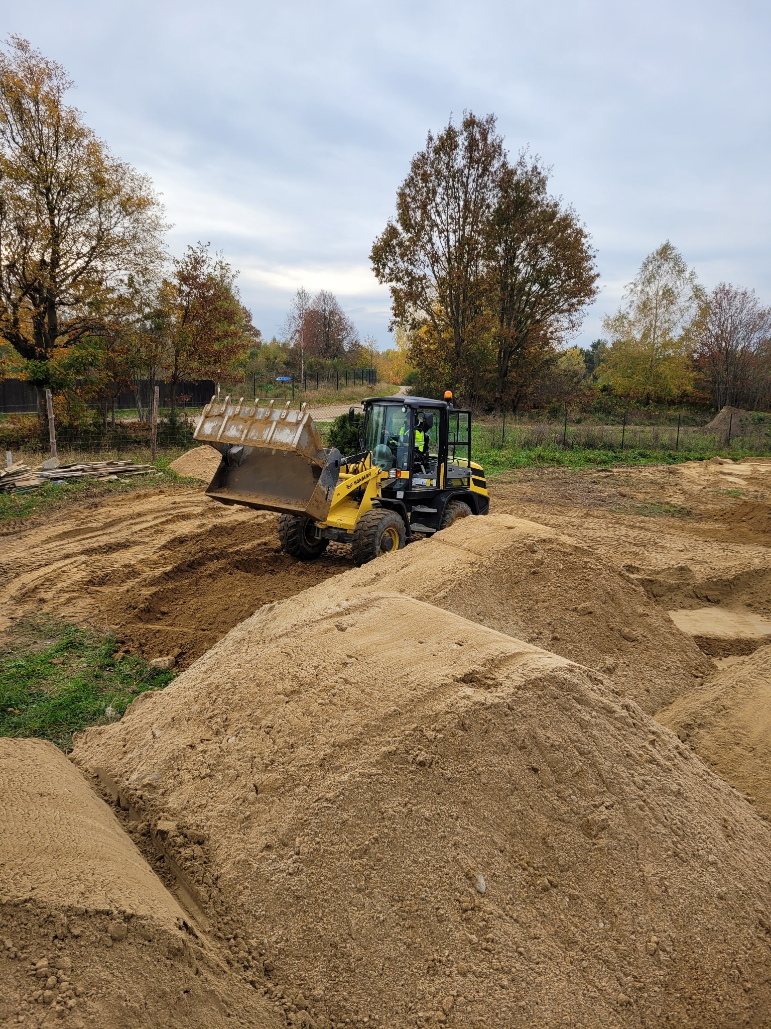 Żółta ładowarka Yanmar na placu budowy, transportująca piasek na tle drzew i pochmurnego nieba. Operator w kamizelce odblaskowej w kabinie.