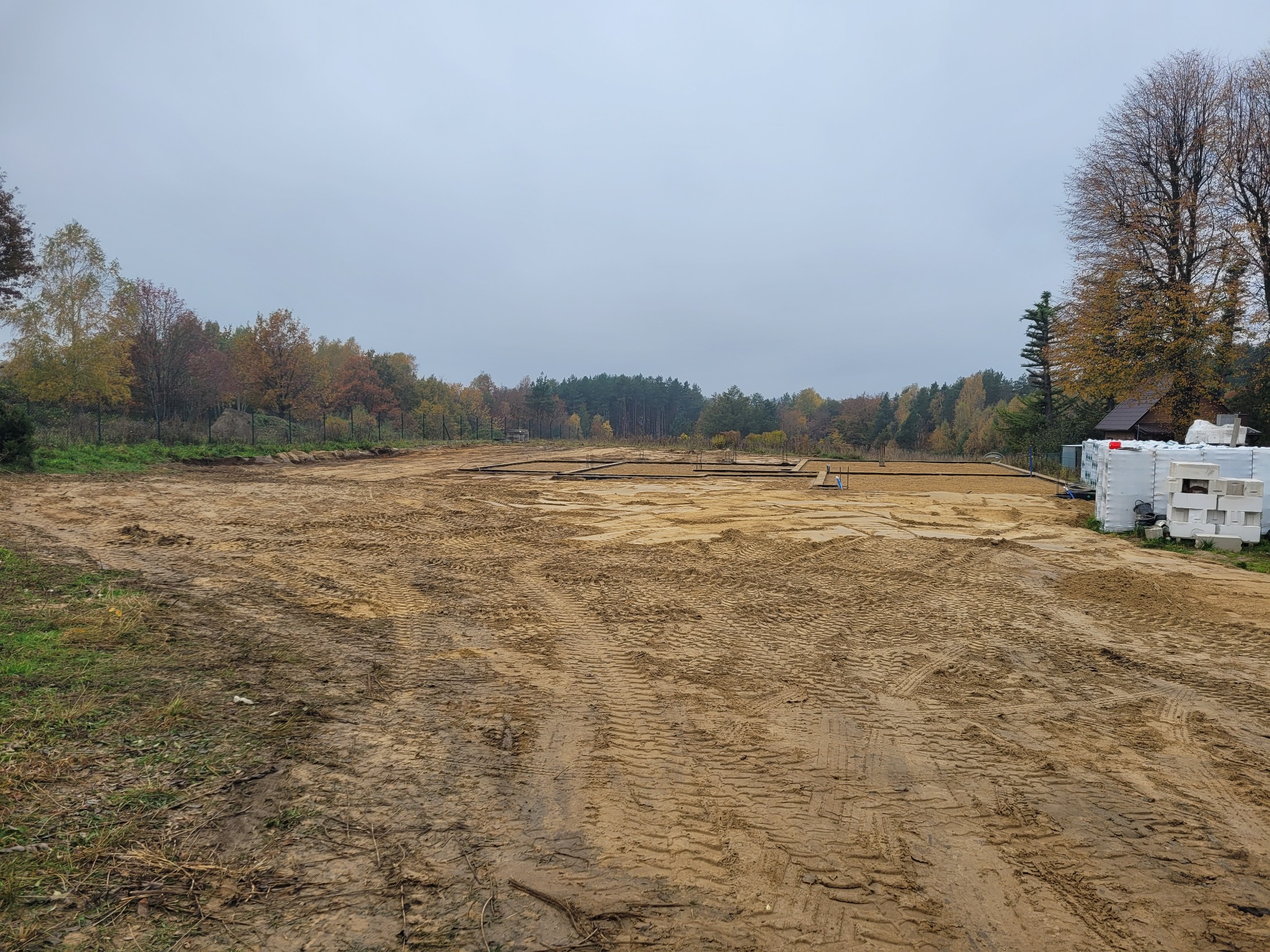 Przygotowany teren pod budowę domu z widocznymi fundamentami i materiałami budowlanymi, jesienny krajobraz w tle. Ślady opon na piaszczystej ziemi.