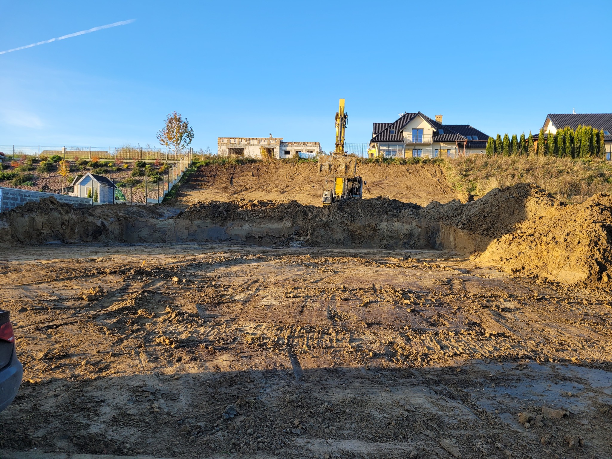 Wykop pod budowę domu jednorodzinnego w słoneczny dzień. Widoczny żółty sprzęt budowlany i ziemia. W tle zabudowa domów i błękitne niebo.