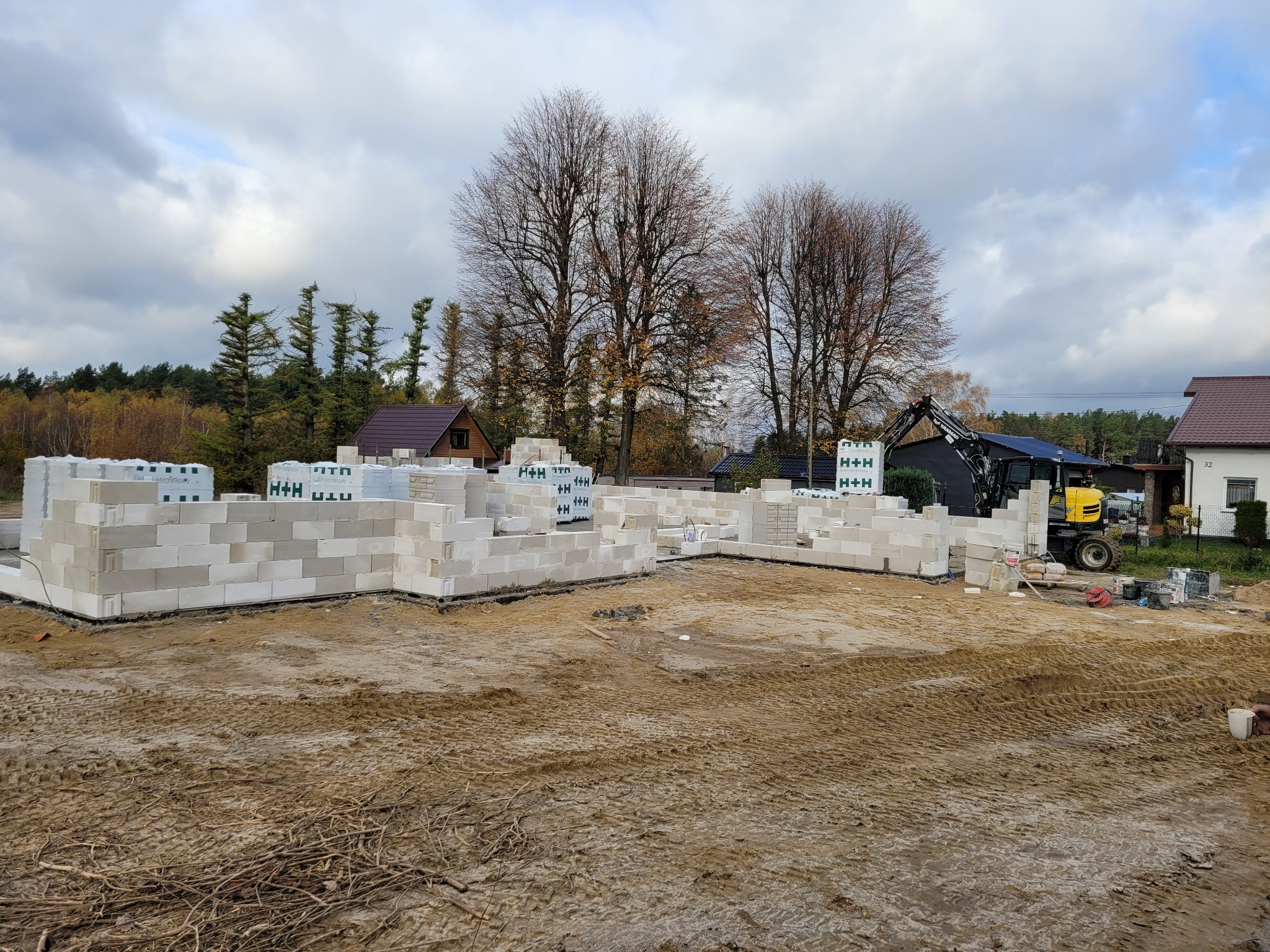 Wznoszenie ścian parteru domu z bloczków H+H. Na paletach składowane bloczki. W tle inne domy i drzewa. Żółta koparka stoi obok budowy. Pochmurne niebo.