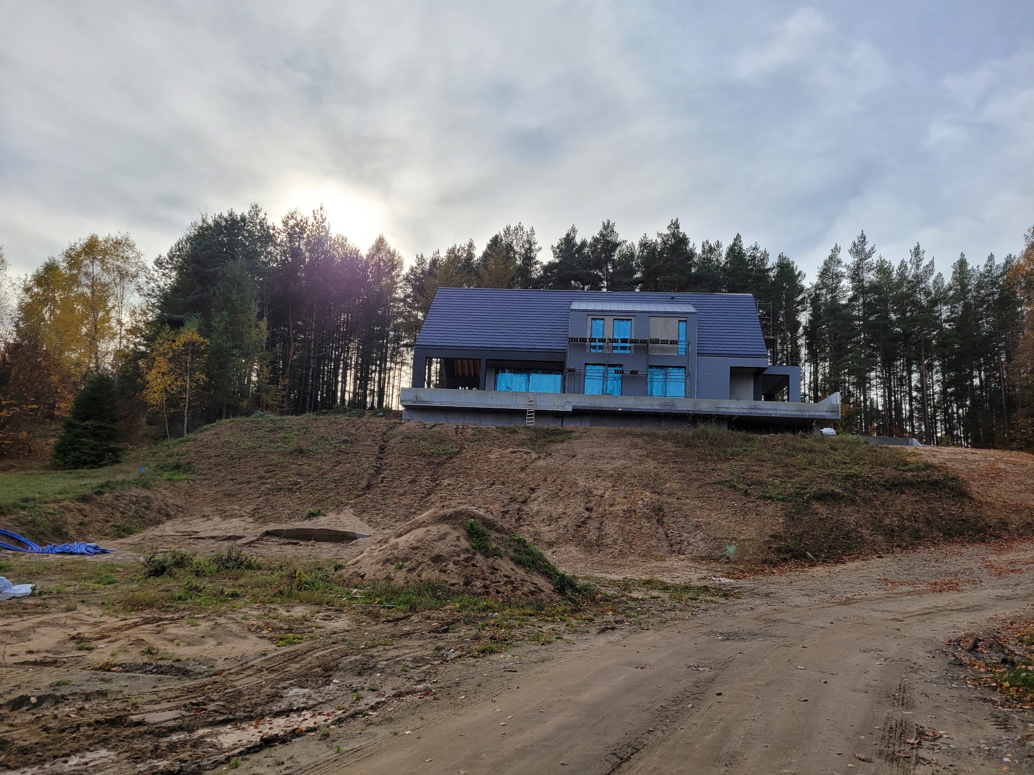 Surowy dom z niebieskimi oknami na wzgórzu, otoczony lasem iglastym w jesiennej scenerii. Widoczny teren budowy i droga dojazdowa.