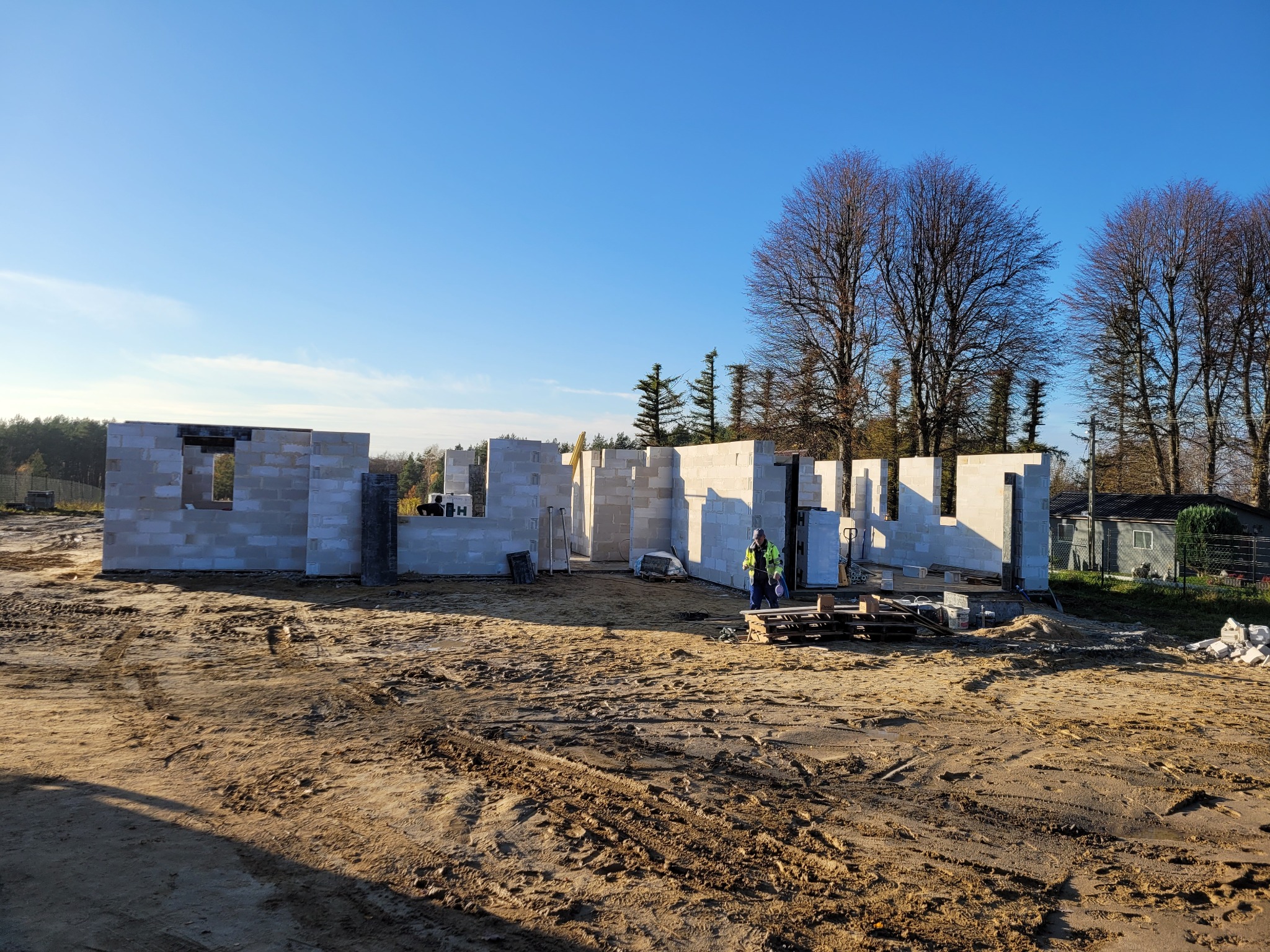 Wznoszenie ścian parteru z betonu komórkowego, widoczny pracownik w odblaskowej kamizelce i palety z bloczkami. Budowa w toku, słoneczny dzień.