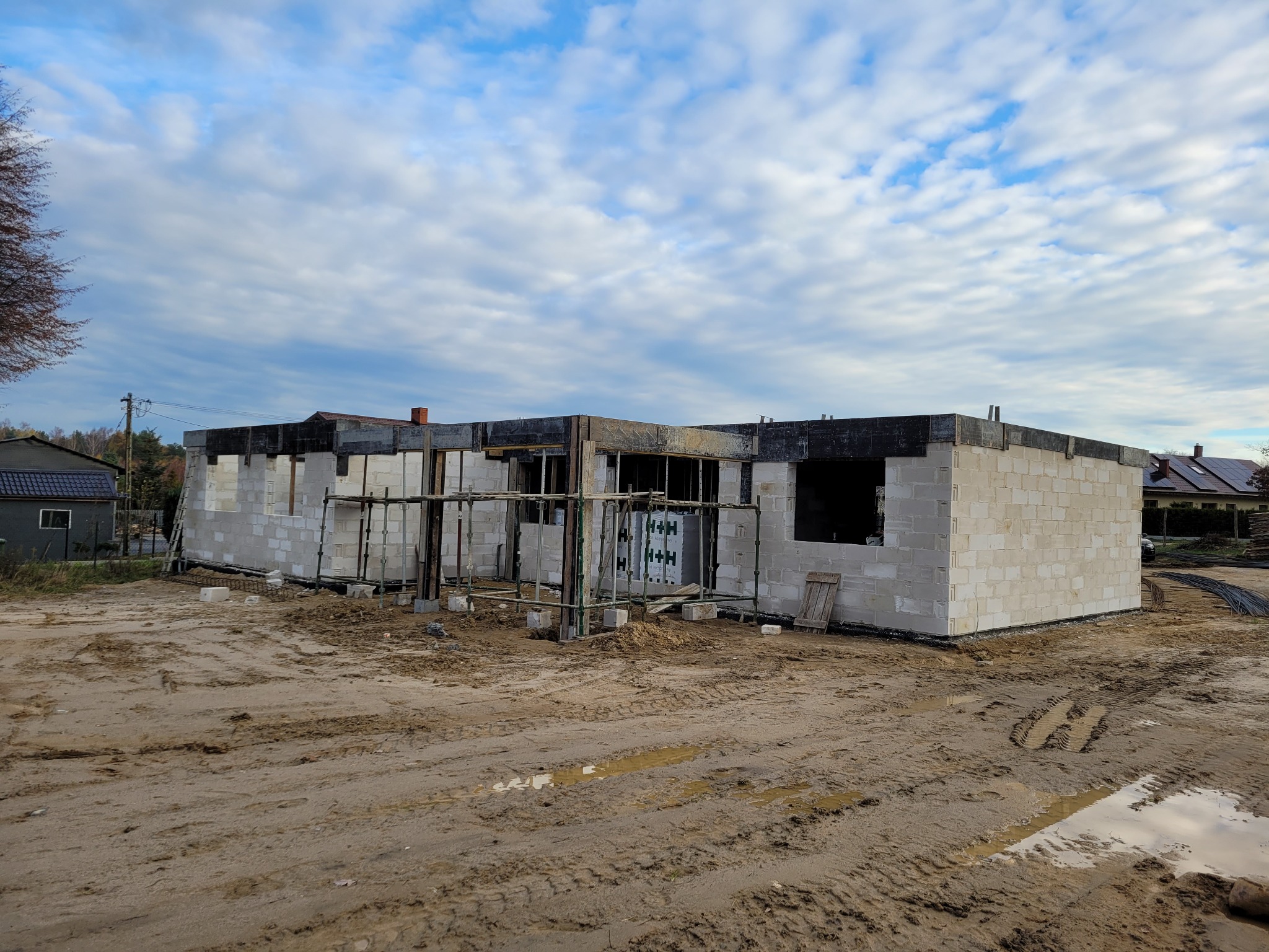 Niedokończony dom z białych bloczków, widać rusztowania, teren wokół błotnisty, pochmurne niebo. Budowa w toku, widoczne detale konstrukcyjne.